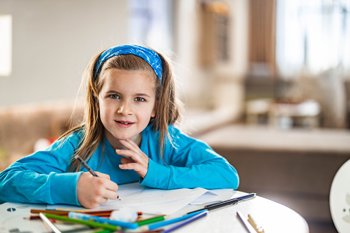 Smiling elementary kid enjoying in coloring on paper at home and looking at camera.