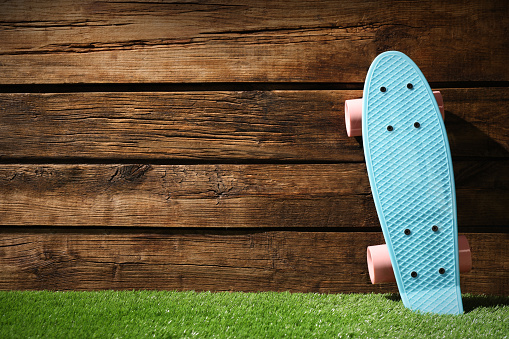 Light blue skateboard with pink wheels on green grass near wooden wall, space for text
