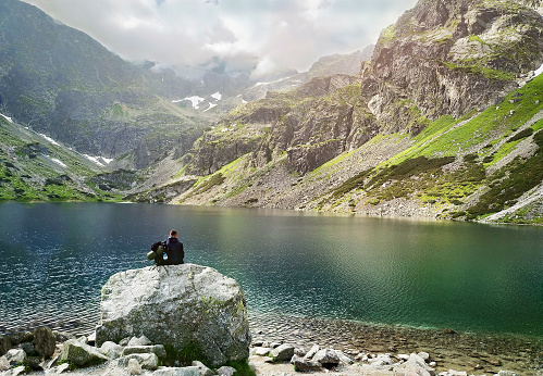 Tourist man sits next to the clear, beautiful, calm lake under wonderful mountains during a tourist trip. Freedom, adventure concept.