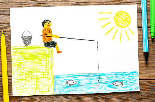 A fisherman with a fishing rod sits on the bank of a river or lake. Children's drawing with felt-tip pens. Wooden table