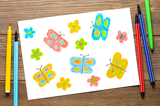Lots of colored butterflies and flowers on a white background. Children's drawing with felt-tip pens. Wooden table.