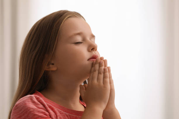 istock Cute little girl with hands clasped together praying on blurred background. Space for text 1393780265