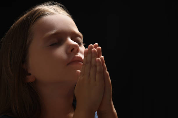 Cute little girl with hands clasped together praying on black background. Space for text
