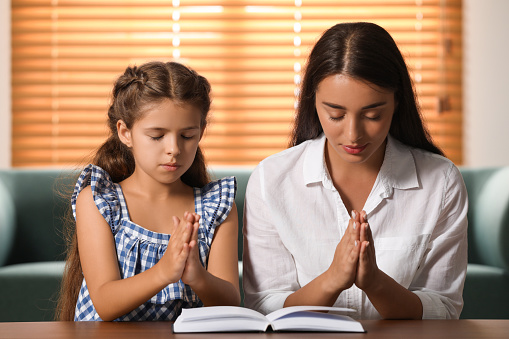 Young woman with her little daughter praying together over Bible at home