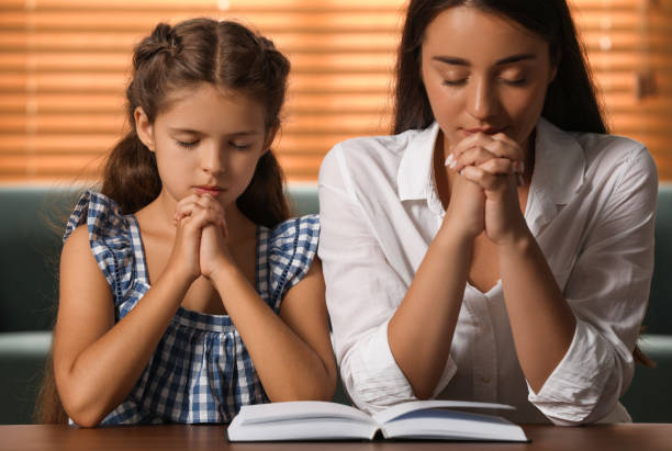 Young woman with her little daughter praying together over Bible at home Young woman with her little daughter praying together over Bible at home praying child christianity family stock pictures, royalty-free photos & images