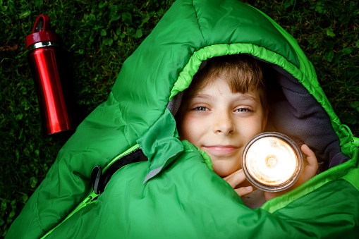 Preteen school kid boy in sleeping bag camping. Outdoors activity with children in summer. Fun and adventure camp, family and friends vacations or weekend trip. Portrait of child with flashlight