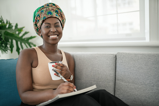 African woman studying from home, writing down notes in book. Relaxing on sofa at home. Looking into camera