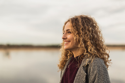 Happy blonde curly beautiful girl in coat against sky. Charming smile. Close up portrait. Spring break. Woman enjoying vacation. Visit local attractions. Girl in nature. Feel happiness.