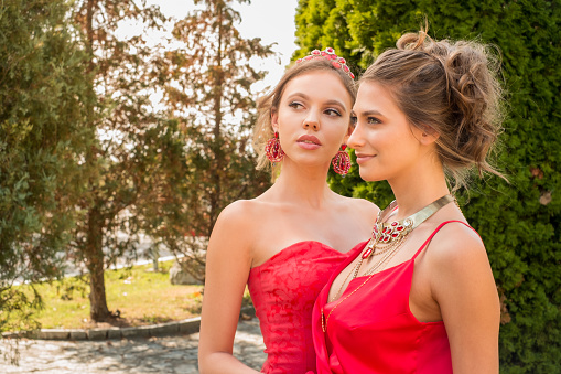 Two beautiful young women in red sleeveless tops wearing ruby jewelry, standing together in public park and looking away, beauty and fashion industry