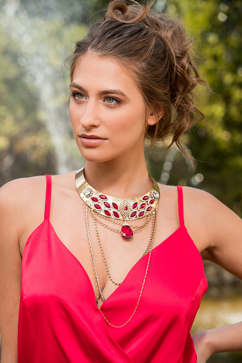Beautiful young woman in red sleeveless top wearing jewelry, standing in public park and looking away, beauty and fashion industry
