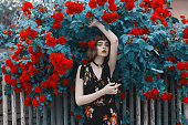 バラの茂みの背景に赤毛のモデル。素晴らしい夏の背景にドレスで赤い唇を持つ素晴らしいアジアの女の子。薔薇園。女性の肖像画。ブルーの調色。シアン色。春の花。ボブの髪型