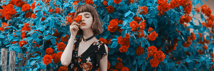 Redhead model on background of roses bush. Fabulous asian girl with red lips in dress on awesome summer background. Rose garden. Woman portrait. Blue toning. Cyan color. Spring flowers. Bob hairstyle