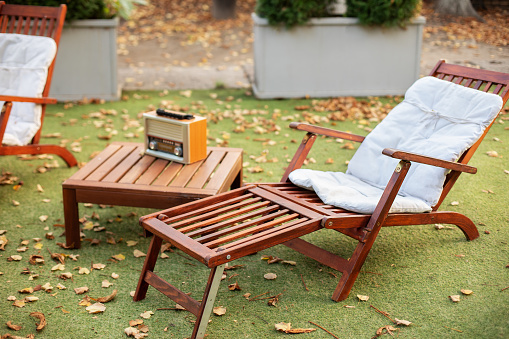 Wooden deck chair on green summer lawn on picnic. Lounge sunbed. Wooden garden furniture on grass lawn outdoor for relax. Backyard exterior. Wooden chair in autumn garden. Vintage radio on table.