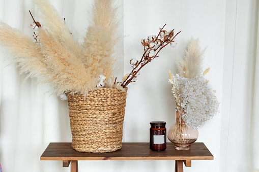 Cozy autumn decor room: on wooden table wicker basket with dried flowers, candle and vase with pampas grass. Vase with bouquet of dried hydrangea, spikelets, ears and plants. living room interior