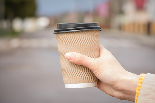 Hand of young woman holding brown paper coffee cup against blurred city background. Mockup image of paper cup for hot beverages, plastic lid. Selective focus