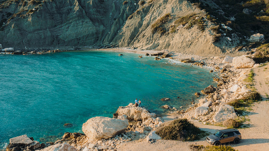 Drone photo of the young heterosexual couple staying on the rock near their car looking at the pure blue sea surrounded by the mountains in South Turkey