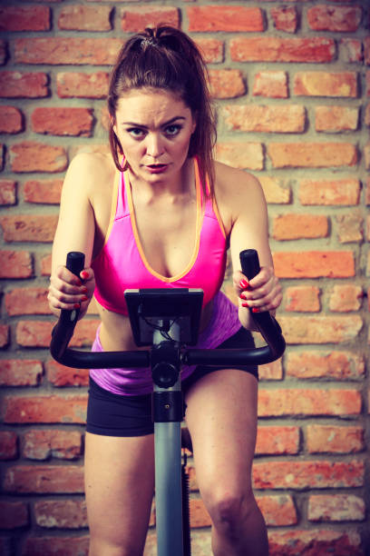 Active woman wearing sport bra working out,  using exercise bike at the gym. Sporty girl training in fitness center. Slim body weight loss concept.