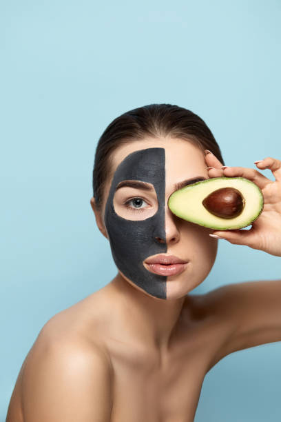 Beauty Facial Mask . Beautiful Young Woman with black mask of clay on face .Skin care .Girl beauty model holding half an avocado in hand . Cosmetic  spa mask . Facial treatment stock photo