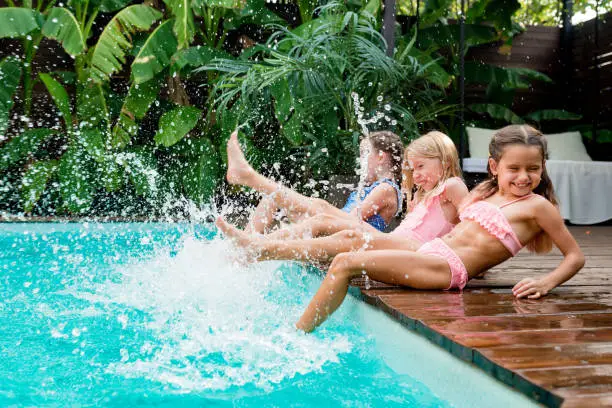 Summertime lifestyle. Girls having fun and relaxing by swimming pool. Kids are sitting at poolside and splashing water with legs on sunny day. Summer outdoors activity for children concept