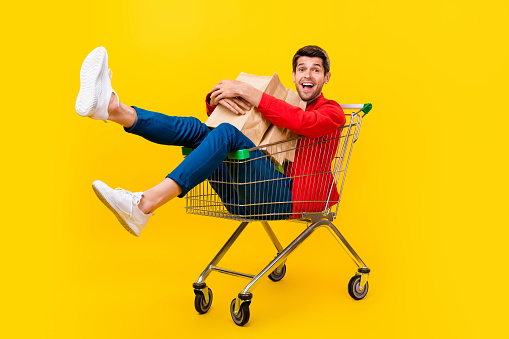 Full length body size view of attractive cheery guy sitting inside cart carrying buyings isolated on bright yellow color background.