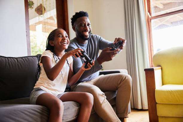 Laughing girl and her dad playing video games together at home Little girl and her father laughing while playing video games together on a sofa in their living room at home video game stock pictures, royalty-free photos & images