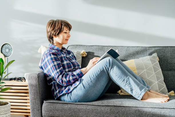 istock Woman sit on the sofa couch and relax while reading a book at home in cozy modern interior living room with green plants. Offline activity lifestyle. Reading hobby. Leisure indoors entertainment. 1393811746