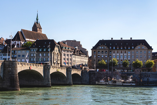 View of the old town of Basel, Switzerland, with an ancient stone bridge over the Rhine river and historical buildings and a church with a high sharp spire, a tourist ship at the pier