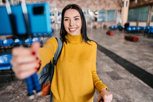 Young female tourist taking a selfie using a phone while waiting for a train or a bus at the station.