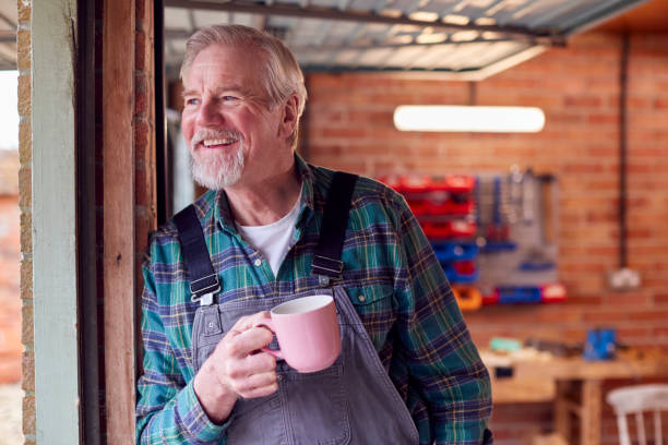 ritratto di un uomo anziano che indossa una tuta nel laboratorio del garage con bevanda calda - stanza degli uomini foto e immagini stock