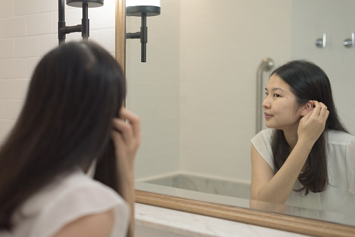 Over the shoulder, medium shot of a cheerful middle aged Asian woman looking at bathroom mirror, checking facial skin condition. Cosmetic, skincare, make up, satisfaction and aging concept