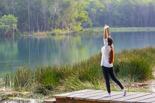 Woman stretching at the pier next to natural lake in the forest for morning exercise and yoga workout routine in the peaceful serene environment