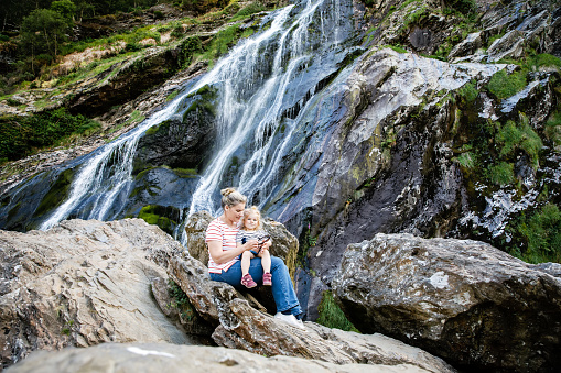 Cute toddler girl and mother sitting near water cascade of Powerscourt Waterfall, the highest waterfall in Ireland in co. Wicklow. Family time vacations with small children. Woman and baby child