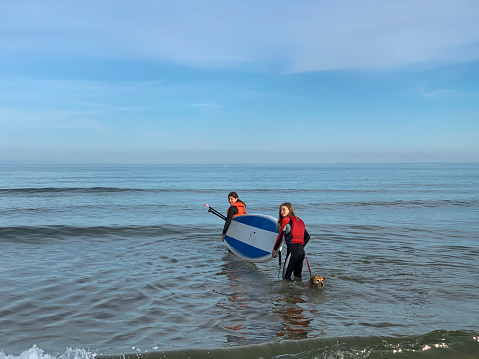 Two teen girls paddle boarding in the sea at a beach in Beadnell, Northumberland. They are wearing wetsuits and are having fun with their pet dog, carrying the board together. They are looking over their shoulder at the camera.