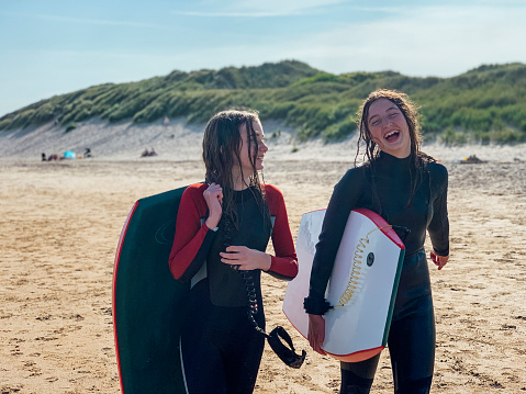 Two teen girls wearing wetsuits holding surf boards under their arms laughing and talking together after a day in the sea at a beach in Beadnell, Northumberland.