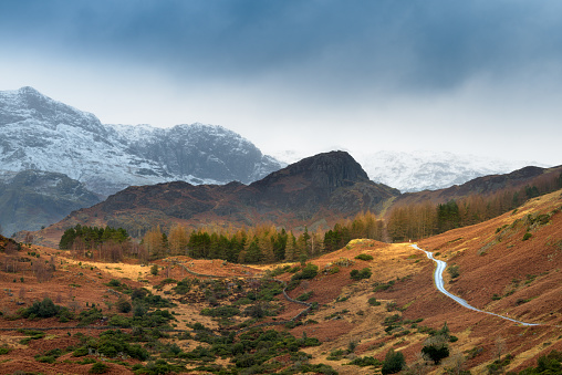 Single track road leading through rural countryside with snowcapped mountains and dark Winter clouds. Langdale, Lake District, UK.