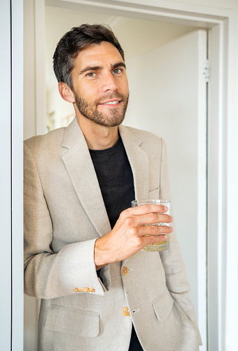 Portrait of a smiling young man wearing a stylish blazer leaning against a doorway at home and holding a drink