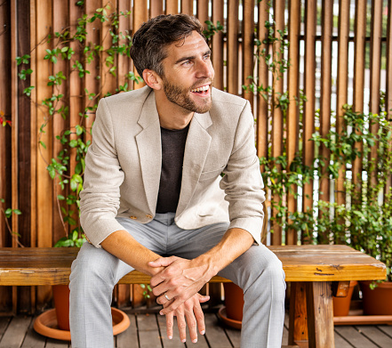 Young man wearing a stylish blazer laughing while sitting on a bench outside on a wood slat patio
