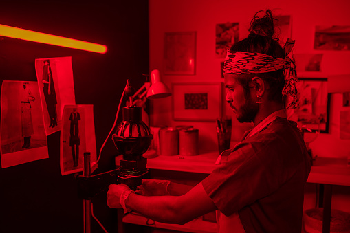 Young man working with film photography in red lit darkroom using photo enlarger to project image onto paper