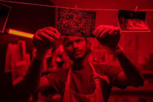 Selective focus shot of young Indian man wearing protective gloves and apron hanging wet photos on thread in darkroom