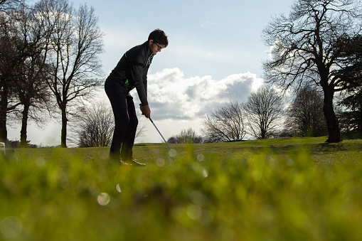 A male golfer is poised to hit the golf ball. Summer day with green grass defocused in foreground, cloudy blue sky and trees around
