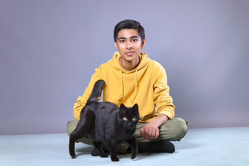 Teenaged youth of Indian heritage wearing a yellow hoodie, playing with his black cat. Focus on youth.