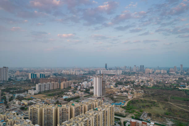 aerial drone shot passing over a building with homes, offices, shopping centers moving towards skyscapers in front of sunset showing the empty outskirts of the city of gurgaon stock photo