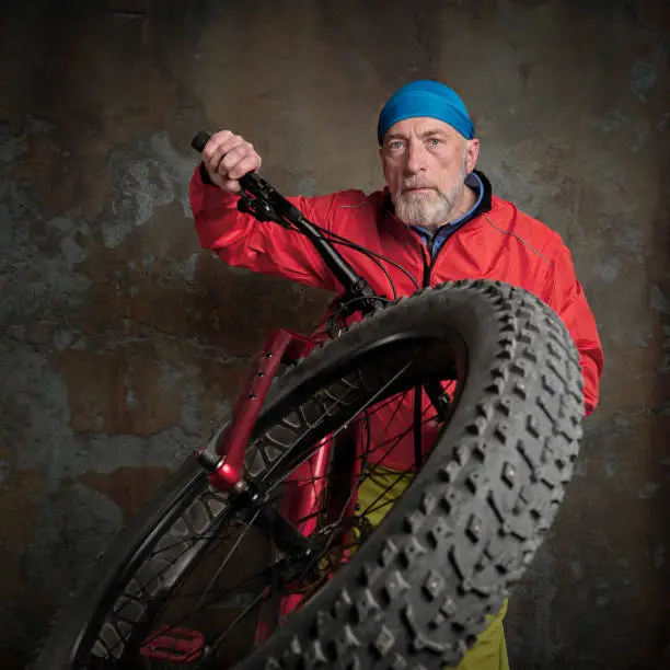 portrait of senior male cyclist in bright red biking jacket and skull cap with a fat mountain bike against a grunge concrete wall