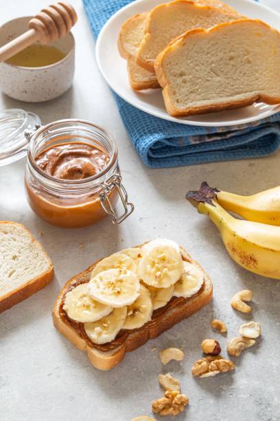 toast avec de savoureux beurre de noix et tranches de banane sur fond clair. noix, noisettes et noix de cajou tartinées dans un bocal en verre. sandwich petit-déjeuner fait maison. concept d’alimentation saine. - appetizer bread breakfast cashew photos et images de collection