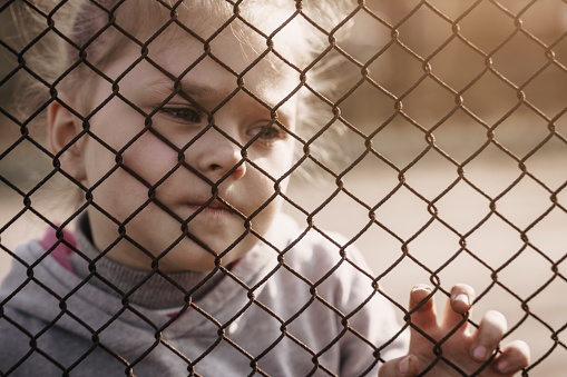 A little girl with a sad look behind a metal fence. The problem of forced deportation of Ukrainian children to Russian-occupied territories.