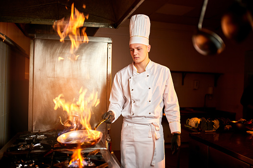 Сhef in the professional kitchen with a frying pan and a fire. Chef's hands hold iron Pan and preparing food on cooker.