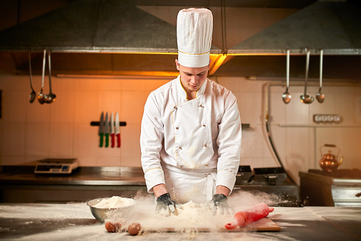 Professional pastry chef prepares dough with flour to make bread, Italian pasta or pizza. Flour is flying in the air on kitchen background. Baking food concept