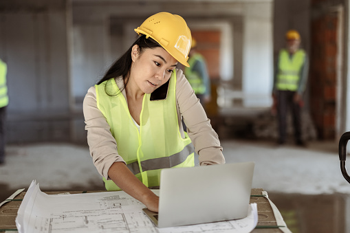Architect with a blueprints using smartphone at a construction site. Portrait of woman constructor wearing white helmet and safety vest outdoors, technology concept