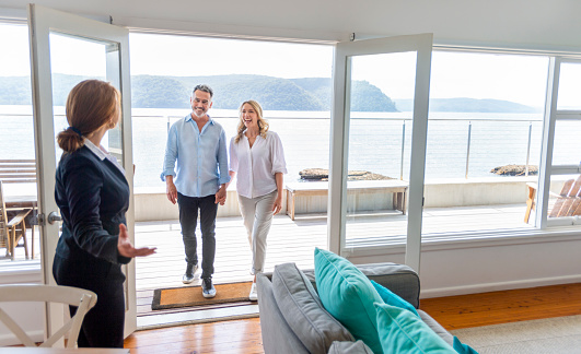 Real estate agent showing a mature couple a new house. The house is contemporary. All are happy and smiling. The couple are casually dressed and the agent is in a suit. Waterfront view can be seen in the background
