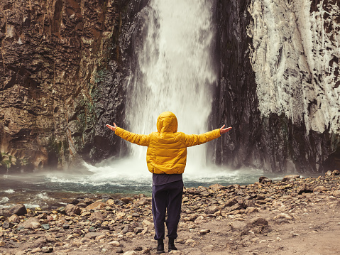 A woman in yellow jacket stands with her hands apart, looking at a stormy ice waterfall flowing down wet rocks.